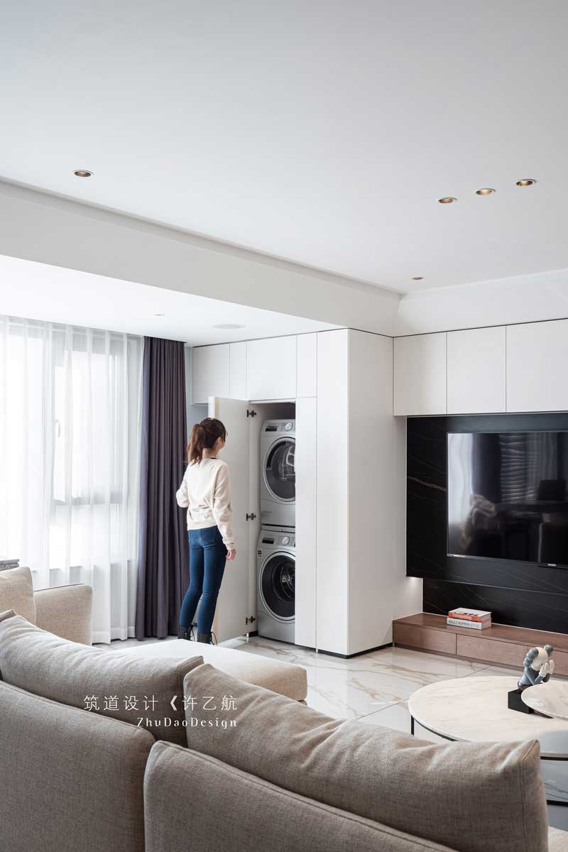 設(shè)計(jì)師充分利用陽臺的空間，把橫在客廳與陽臺的墻體打通，讓整個(gè)陽光通過大面積的窗戶照射進(jìn)來，使整個(gè)空間采光通透；又將電視背景墻與之結(jié)合，并做了隱形門處理，將洗衣機(jī)、烘干機(jī)嵌入，既美觀又實(shí)用。